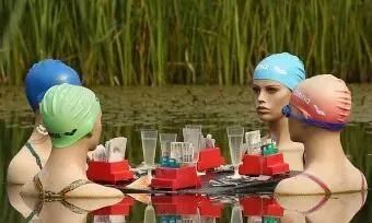 Four mannequin wearing swim caps floating on a calm pond